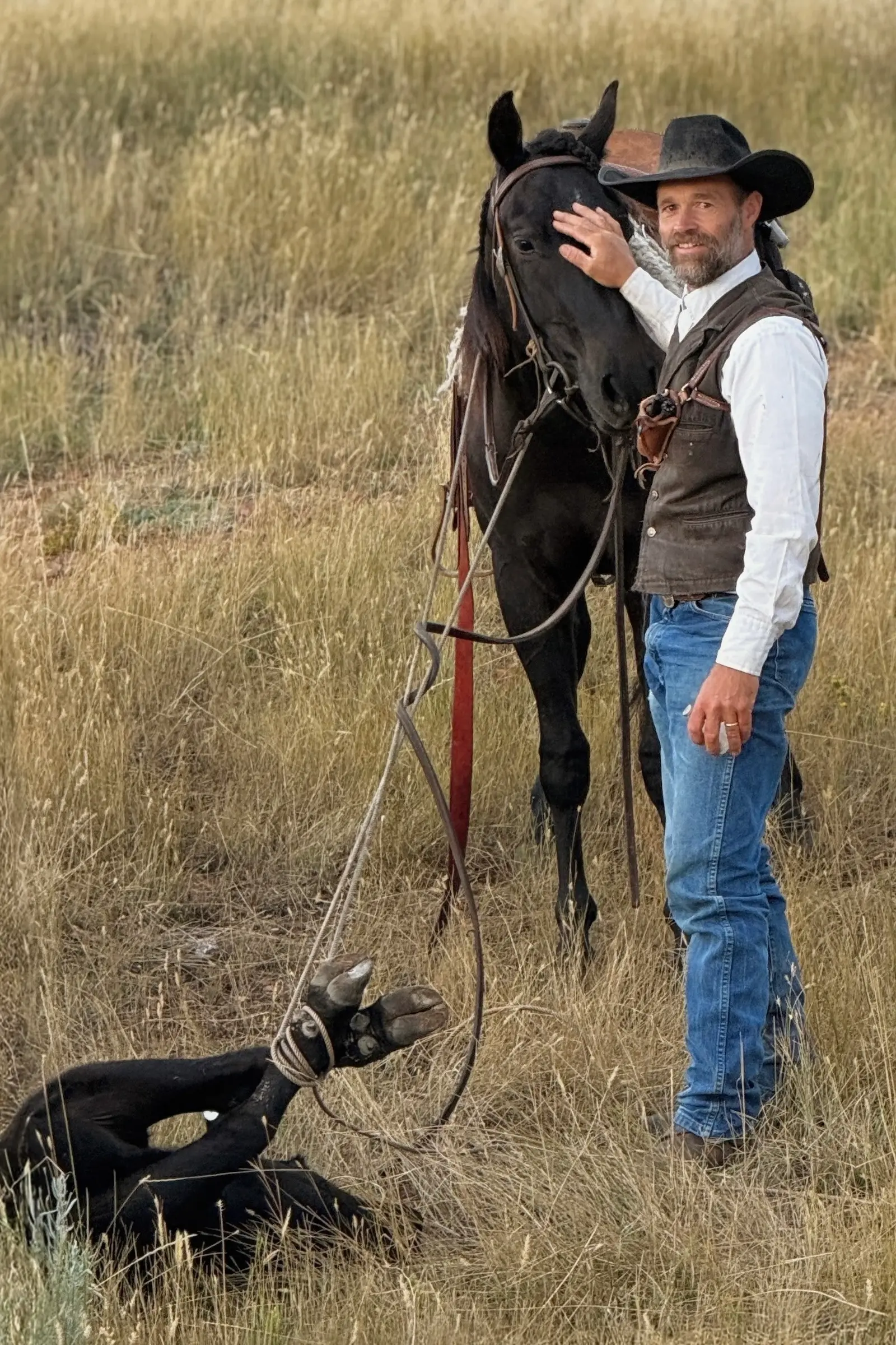Paul Van Dyke in a black hat and leather vest, one hand on his black horse's forehead, with a roped calf tied down in the grass beside them