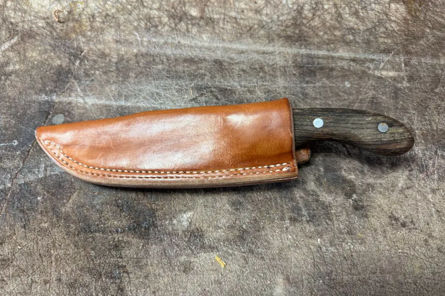 A stitched russet leather knife sheath holding a wood-handled knife, on a worn workbench