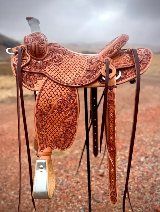 Full custom saddle with basket-stamped seat and fenders and floral borders, standing outdoors on red dirt under a grey sky