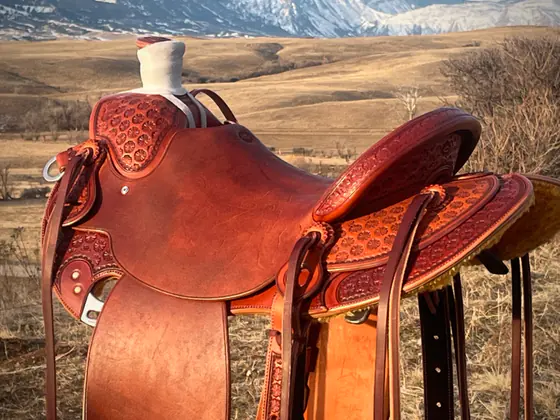 Oiled custom saddle with geometric-stamped swells and skirts, on a stand in the grass with the snow-topped Bighorn Mountains behind