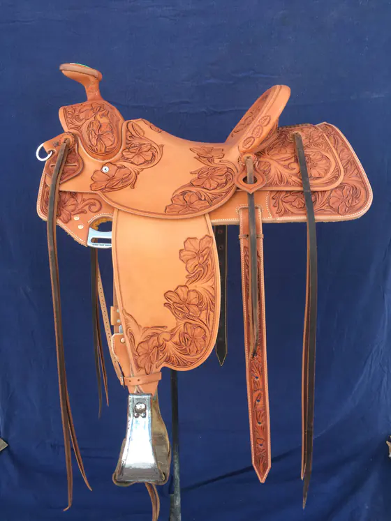 Light veg-tan custom saddle with carved floral corners, swells and fenders, against a blue backdrop