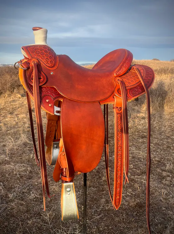 Dark-oiled custom saddle with stamped swells and skirts, on a stand in winter prairie grass