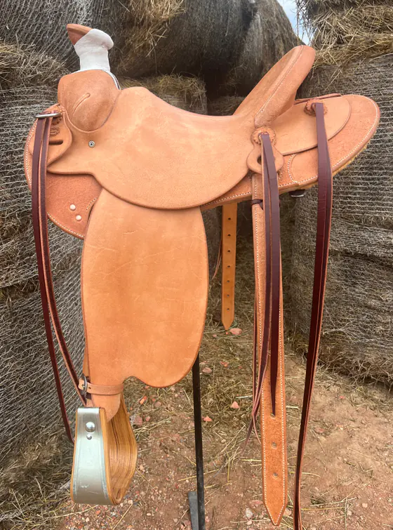 Plain roughout custom saddle with long saddle strings, standing in front of hay bales