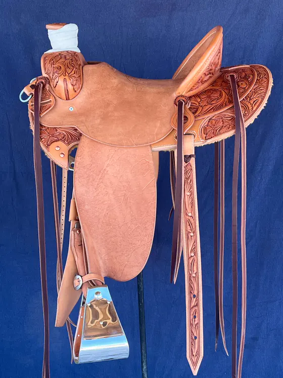 Custom saddle with a roughout seat and fenders and floral-carved swells, skirts and stirrup leathers, against a blue backdrop