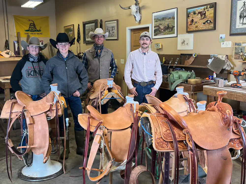 Four students standing in the Sheridan shop behind the saddles they built