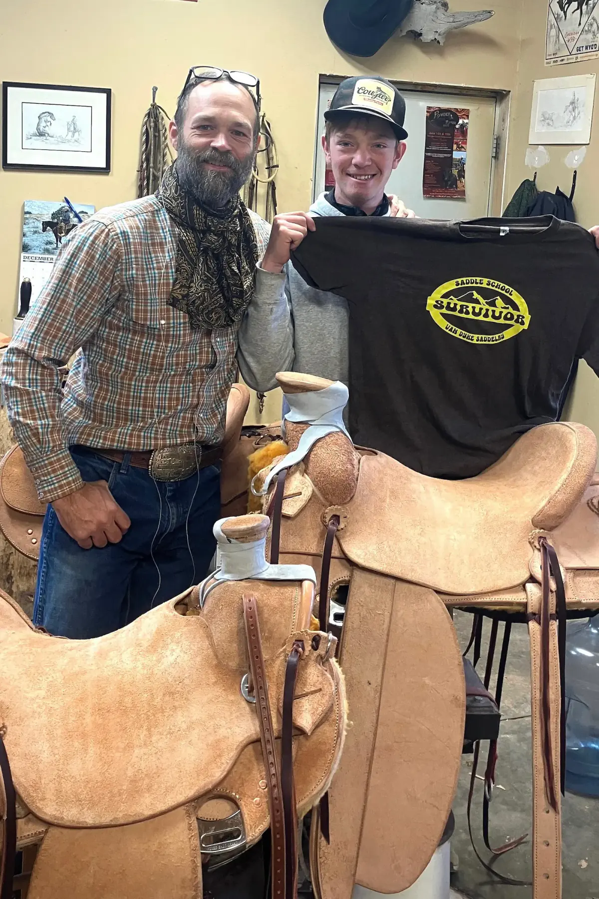 Paul Van Dyke in the Sheridan shop beside a student holding up a Saddle School Survivor t-shirt, with the saddles they built in front of them